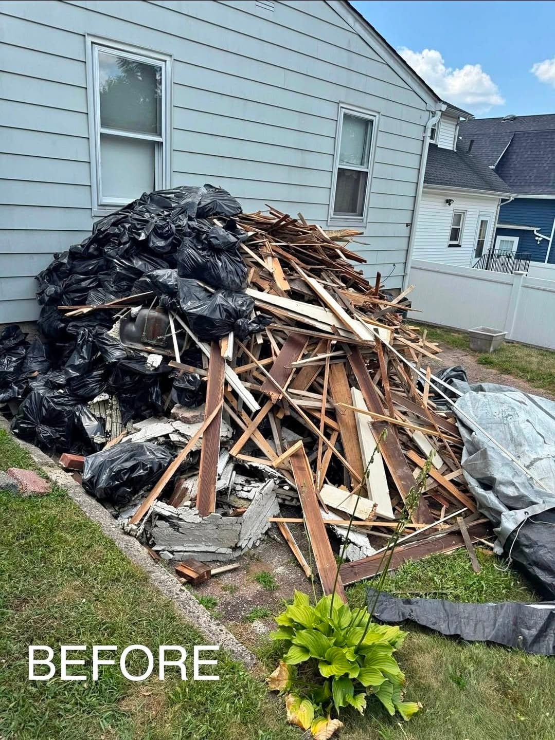 Side yard with massive pile of construction debris including broken wood, boards, concrete, and garbage bags next to house