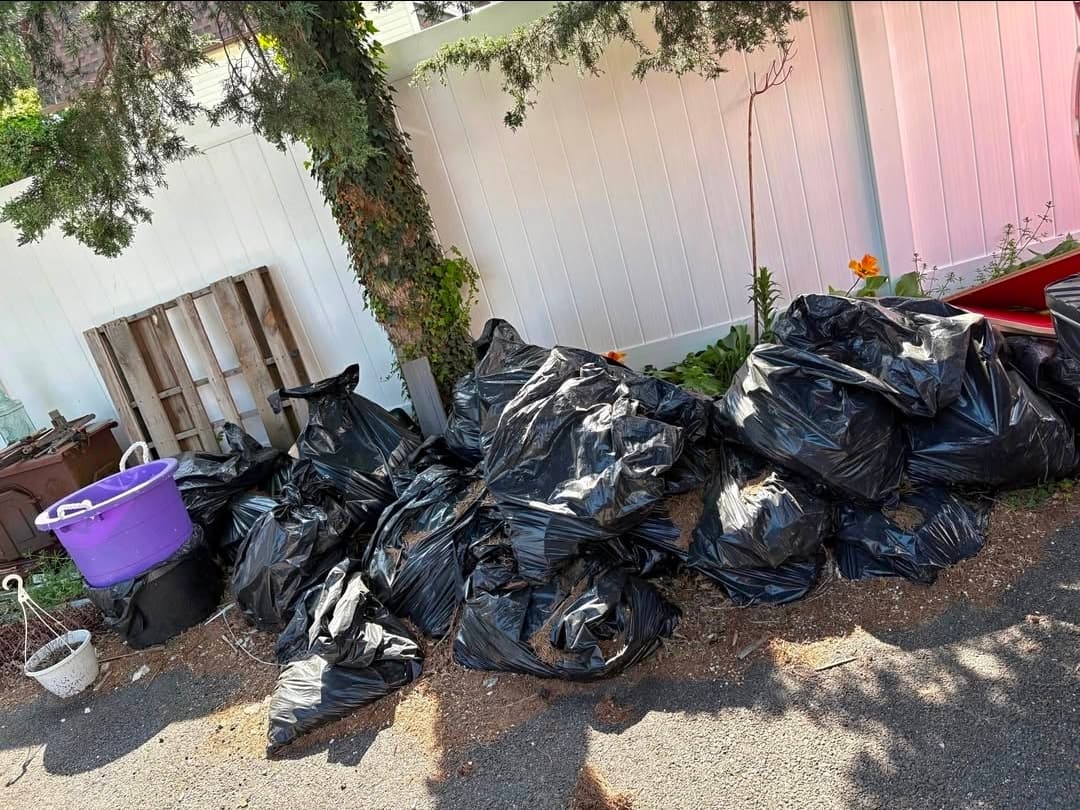 Numerous black garbage bags and household items piled along white vinyl fence